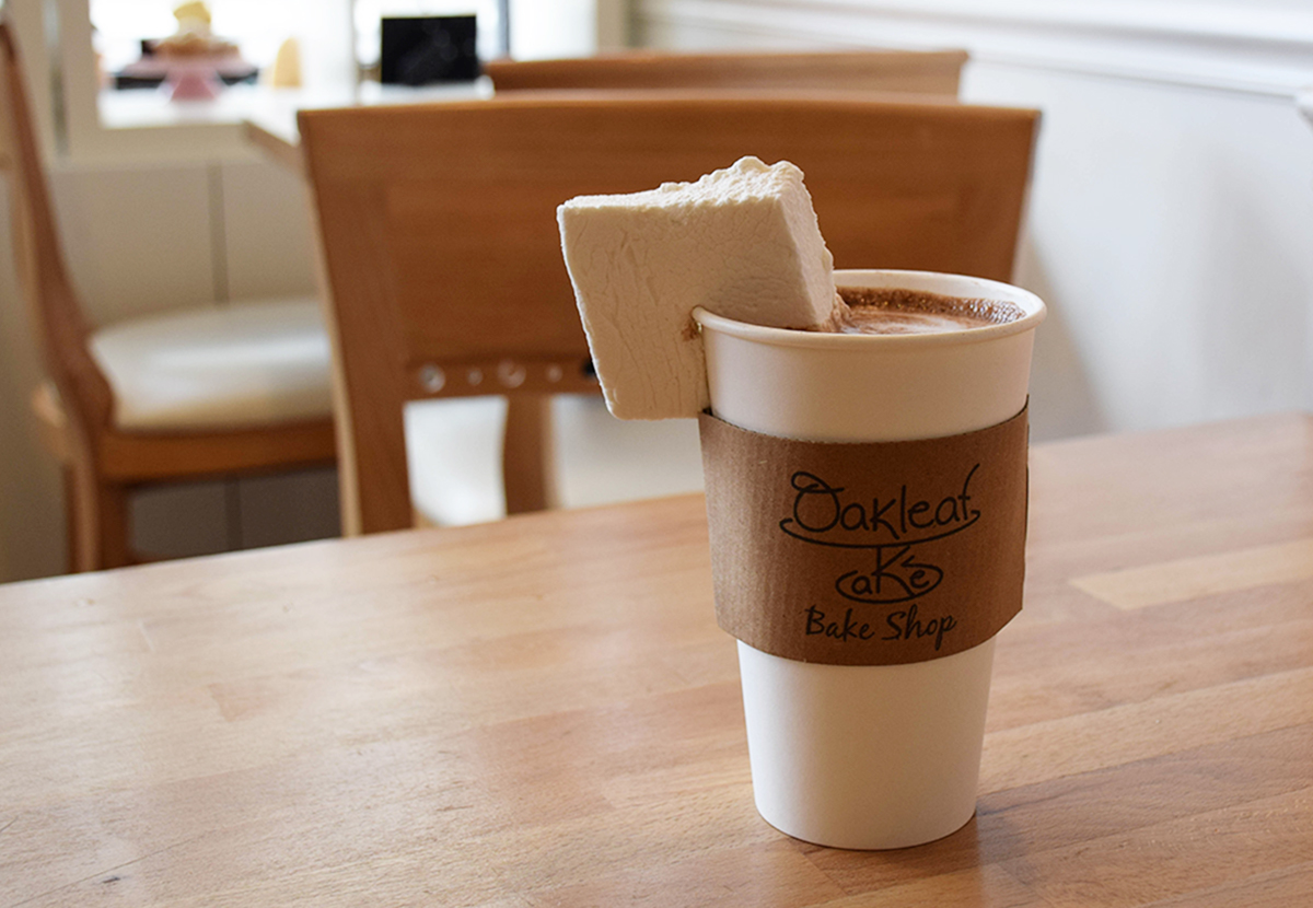 A paper cup of hot chocolate sits on a wooden table, garnished with a giant square marshmallow.