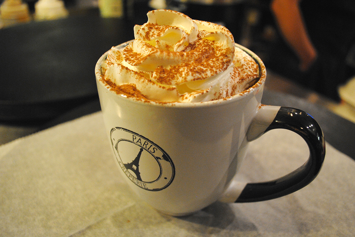 A mug, emblazoned with a restaurant's logo featuring the Eiffel Tower, holds hot chocolate topped with whipped cream.