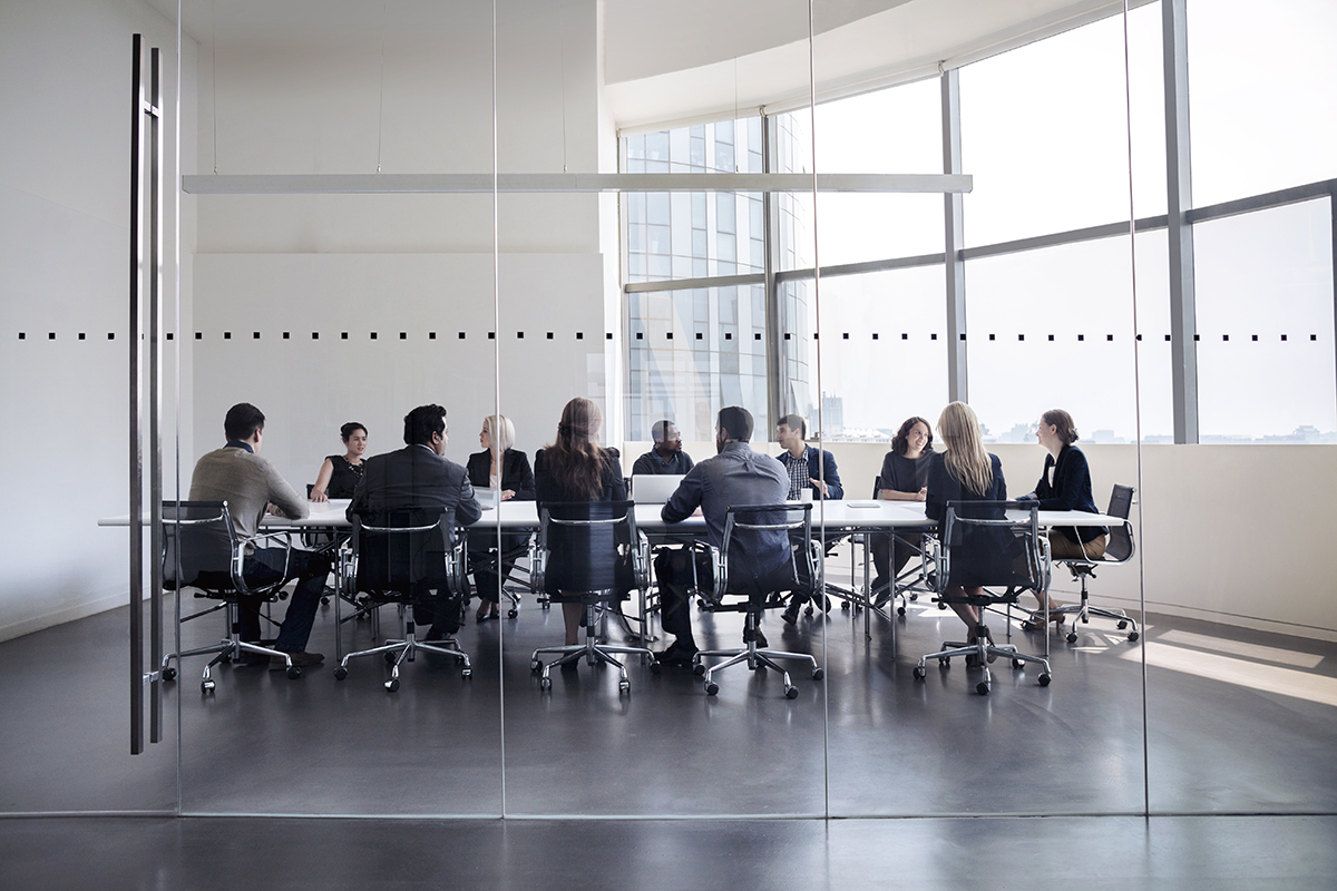 Men and women in suits sit at an office boardroom table