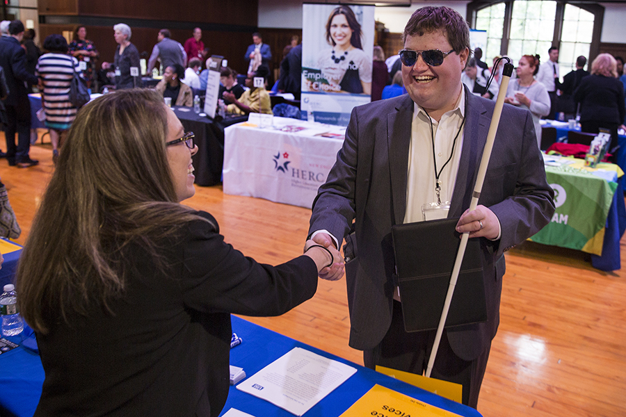 A man wearing sunglasses and holding a white cane shakes hands with a job recruiter