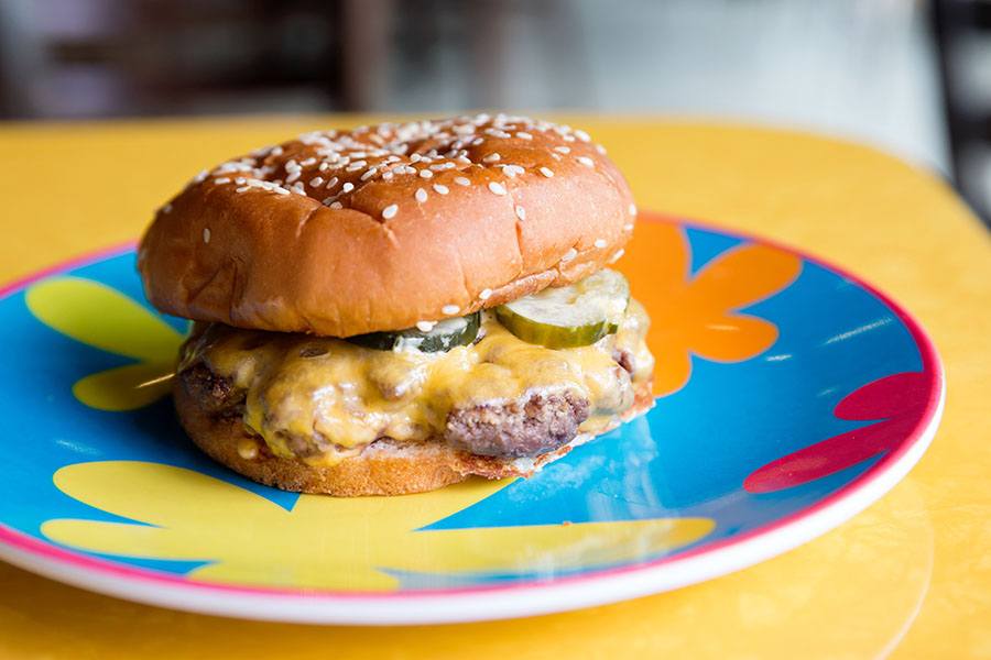 A cheeseburger with pickles sits on a colorful floral plate on a yellow table.