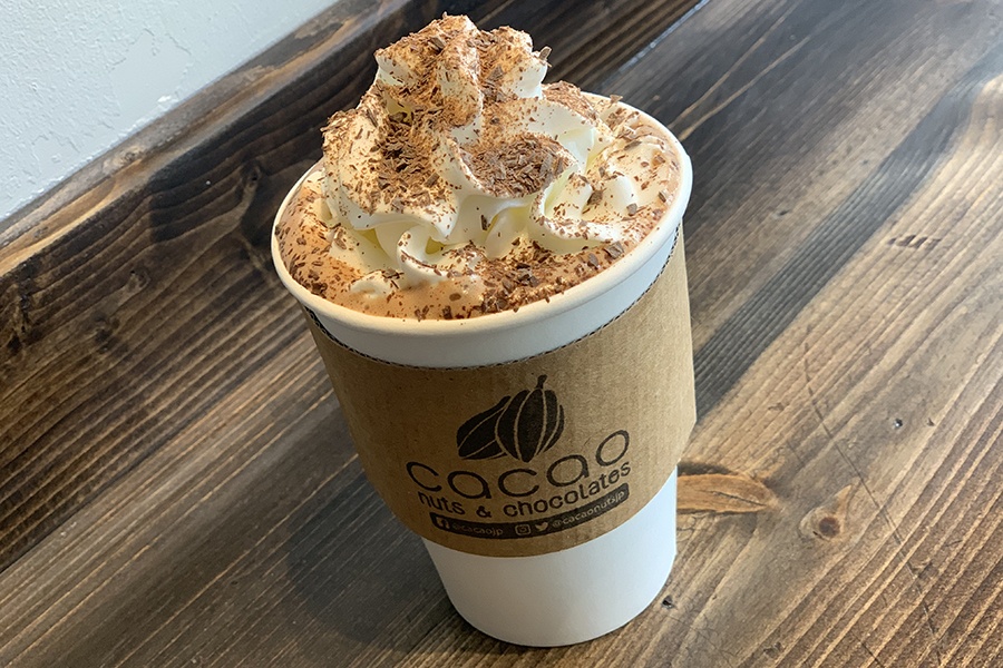 White paper cup and cardboard sleeve holds hot chocolate, topped with ample whipped cream and shaved chocolate.