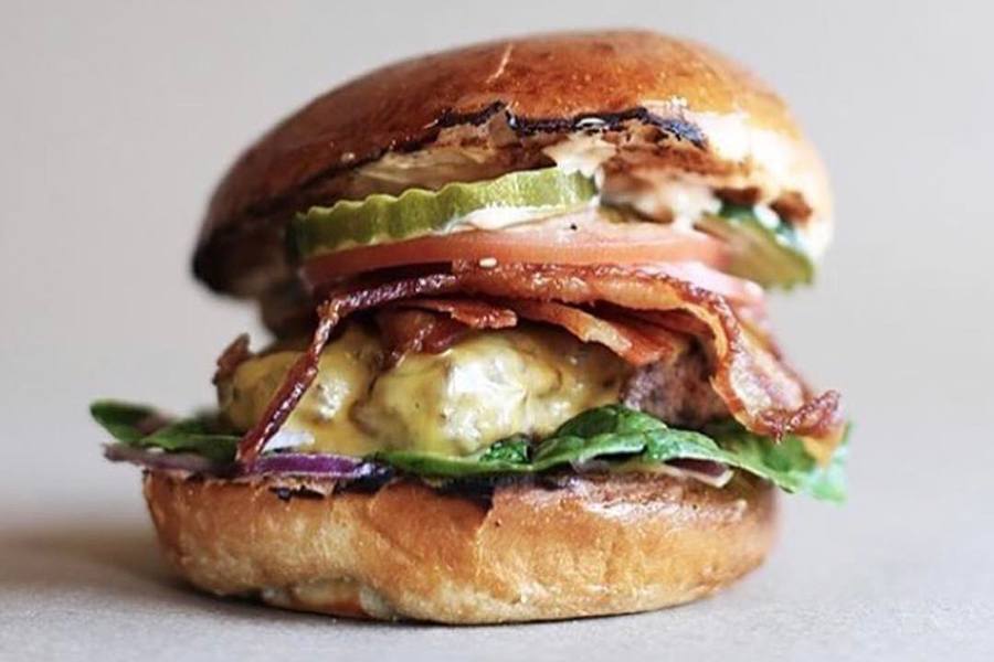 A burger stacked high with toppings like pickles, bacon, and cheese is isolated on a white background.