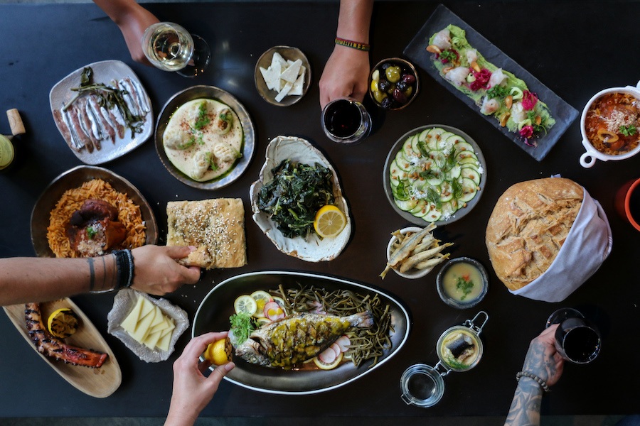 Overhead view of a table full of Green food and wine, with diners' hands reaching in to grab glasses and food.