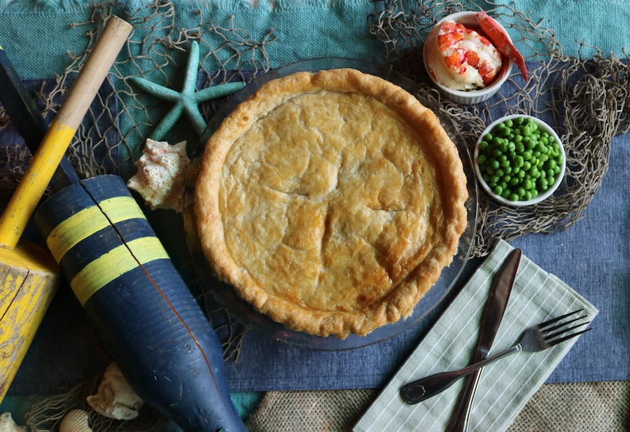 Overhead view of a pie with bowls of peas and lobster to the side and nautical decor elements around it.