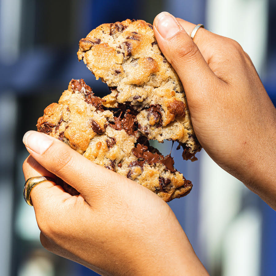 Two hands break a melty chocolate chip cookie in half.