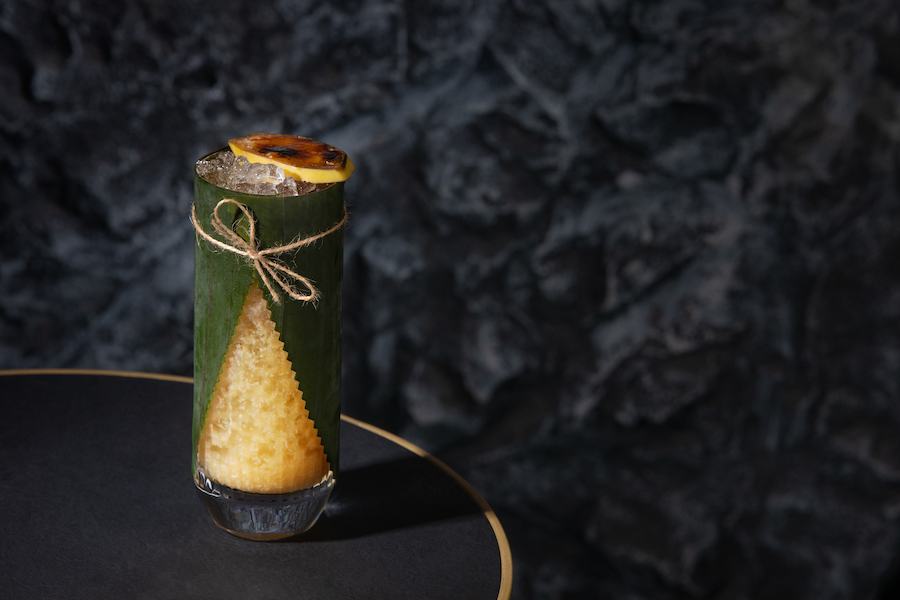 An opaque yellow cocktail is garnished with a burnt citrus slice and served in a tall glass wrapped in a banana leaf. 