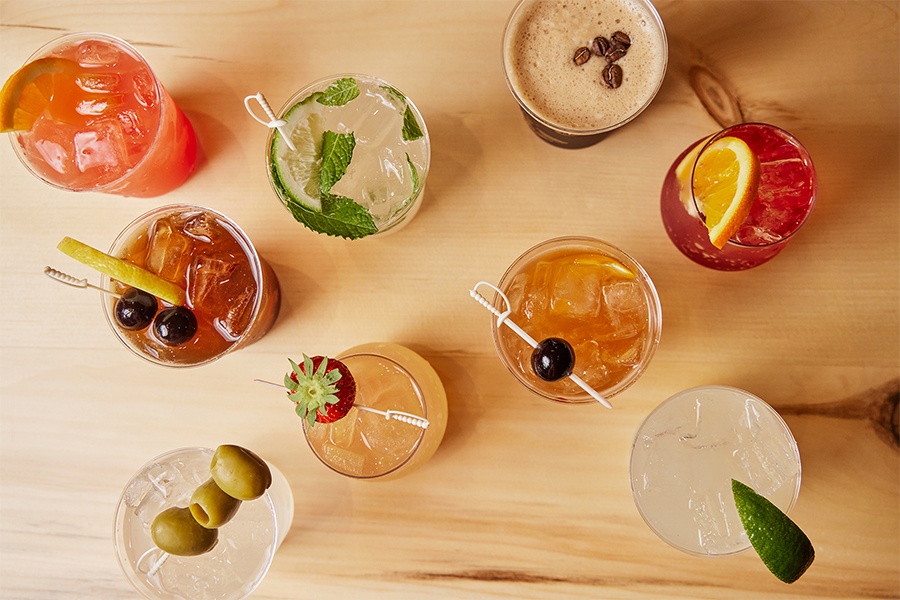 Overhead view of nine different cocktails on a light wooden surface.