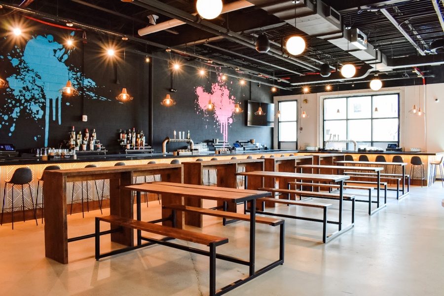 An empty bar features a black wall with bright blue and pink paint splatters, plus long wooden tables with attached benches.