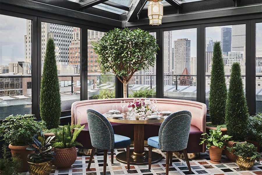 Interior of a greenhouse-style rooftop restaurant space with a decorative tree, a colorful tiled floor, and a pale pink round banquette.