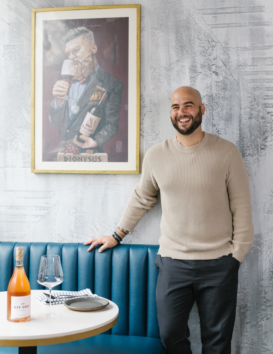 A smiling, bearded man in a long-sleeved beige shirt and gray slacks leans against a teal booth in a restaurant.