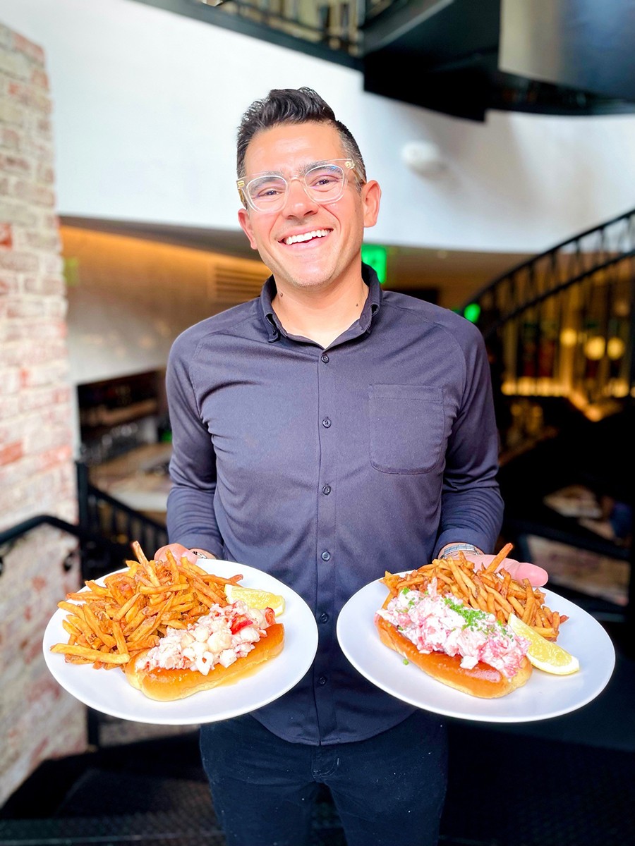 A man stands in a restaurant and holds two plates, with a different style of lobster roll on each.