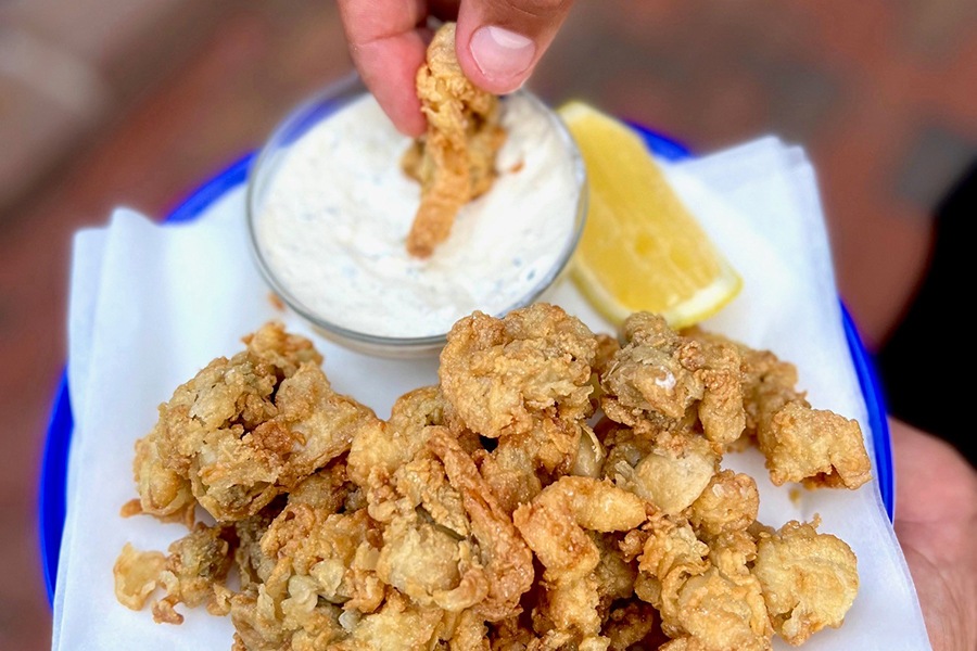 A hand holds up a plate of fried whole belly clams, while another hand dips one clam into tartar sauce.