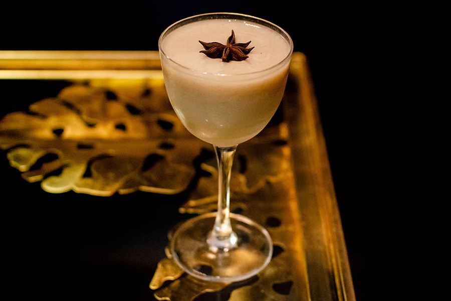A foamy off-white cocktail in a delicate glass is garnished with star anise and sits on a table with golden details.