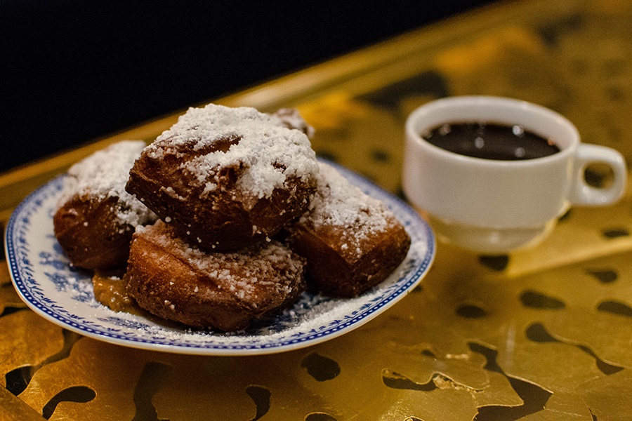Fried balls of dough are topped with copious powdered sugar. A side of chocolate sauce sits nearby.