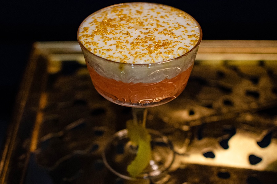 A pink cocktail is topped with foam and golden glitter, and a lemon peel is tied around the glass stem. It sits on a golden table.