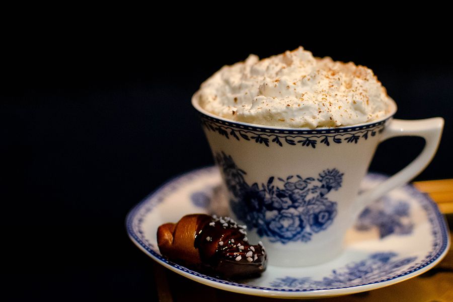 A white teacup and saucer with a delicate blue pattern is full of a beverage topped with thick whipped cream, with a chocolate pretzel nugget on the side.