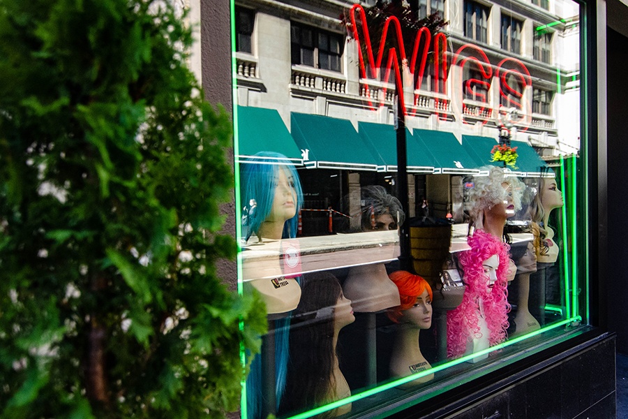 A store window is full of mannequin heads wearing wigs and neon signage that reads "wigs."