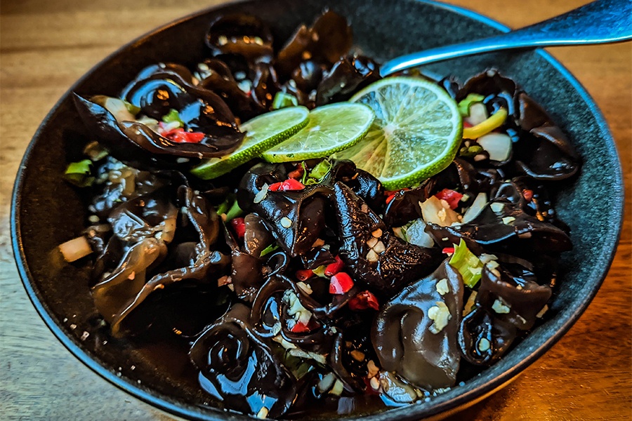 A big bowl is full of shiny black mushrooms, garnished with chopped red chile peppers and thin lime slices.