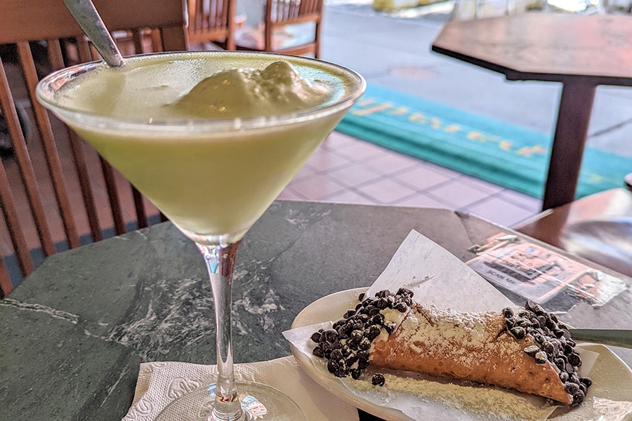 An opaque pale green martini has a scoop of ice cream in the same color, and there's a mini cannoli on the side with chocolate chips.