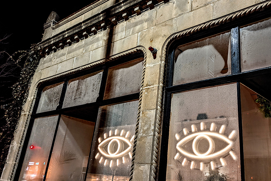 White neon signage that looks like two eyes hangs in the large front windows of a bar, photographed on a rainy night.