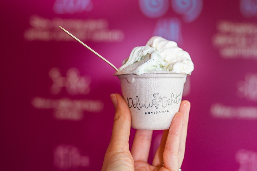 A hand holds up a paper cup of white gelato in front of a pink background.