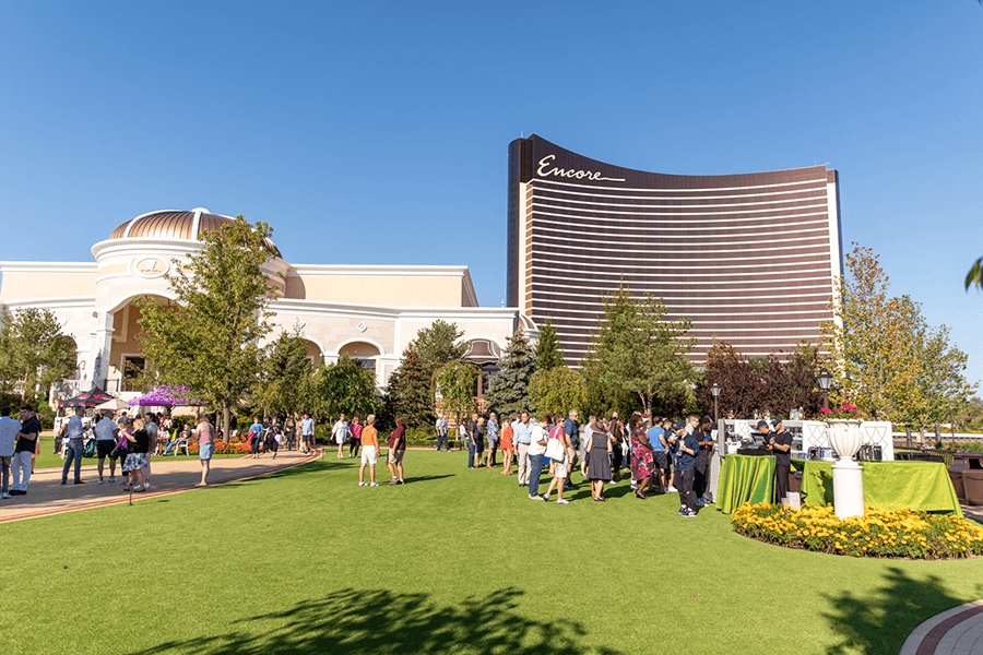 Daytime view of a large green lawn in front of a gold high-rise with Encore written at the top. People are gathered on the lawn for an event.