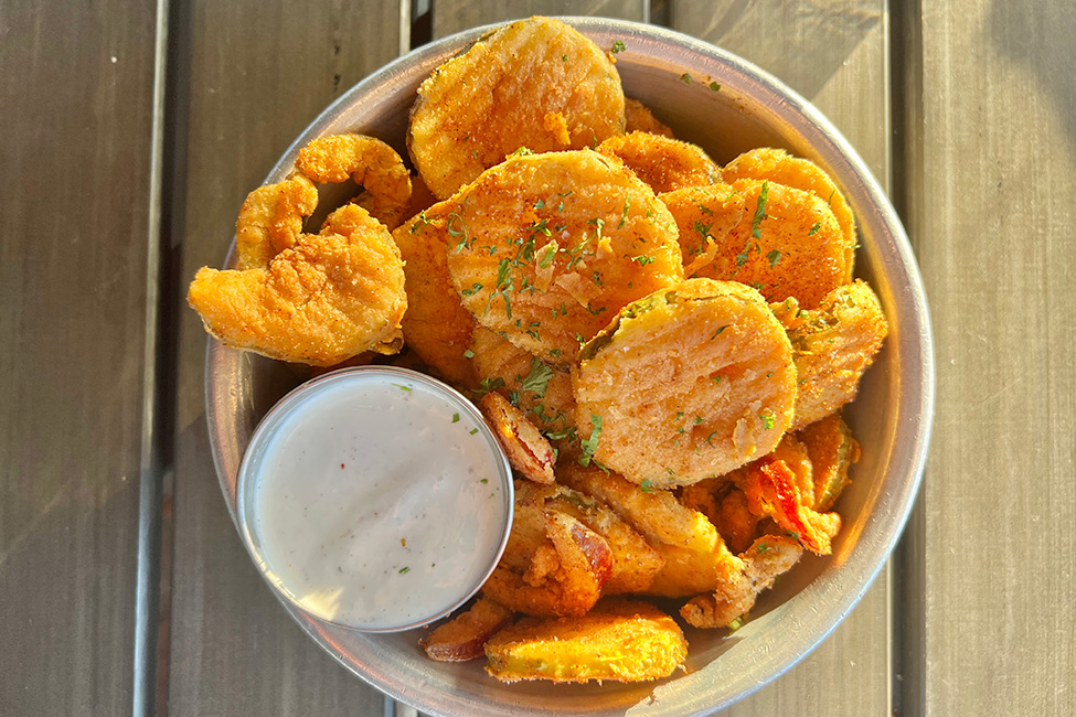 Ridged circles of golden-brown fried pickles are in a light bowl with a small metal bowl of thick, white sauce.