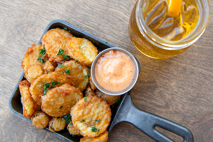 Golden-brown fried pickle rounds are served with thick orange dipping sauce in a small cast-iron pan, with beer on the side.