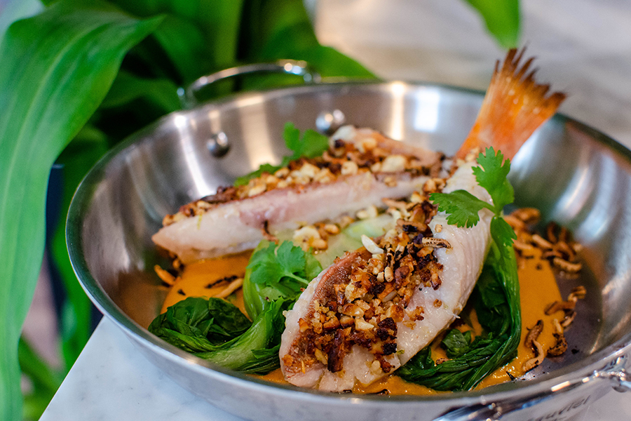 Two pieces of fish with the tails on are garnished with crushed nuts and sit on greens in a red curry-style broth.