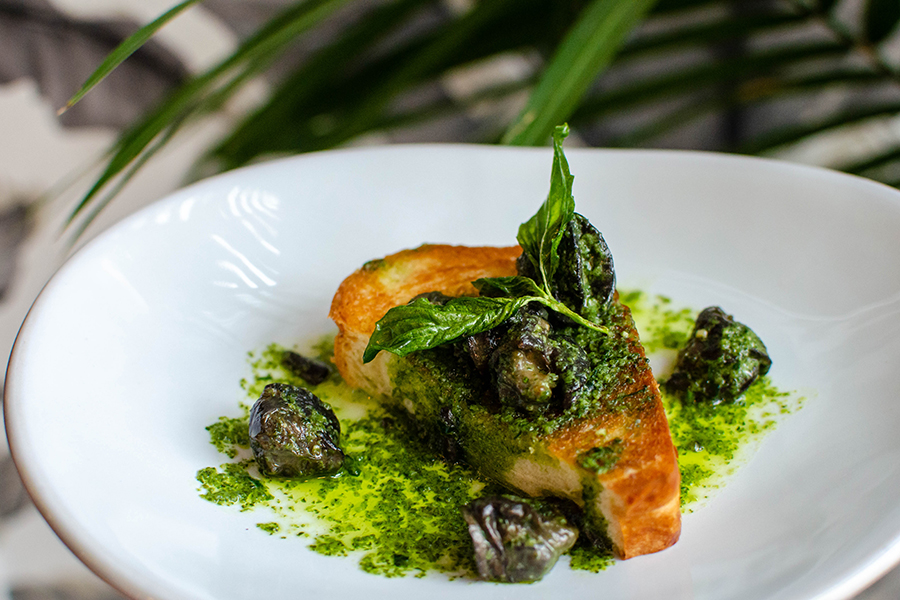 Several plump dark snails sit atop a golden brown piece of bread in a pool of herby butter.