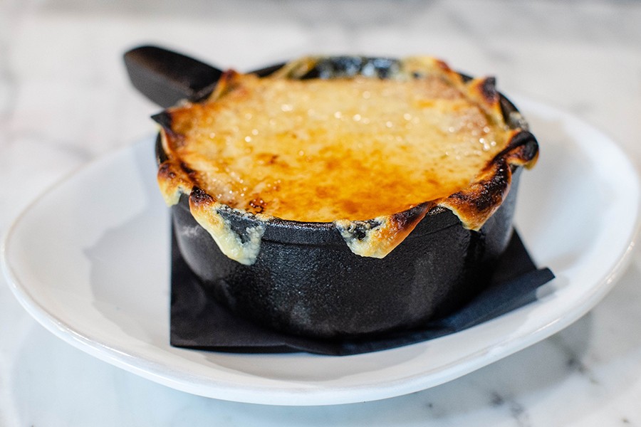 French onion soup is served in a small black cast iron saucepan with charred cheese hanging over the edge.