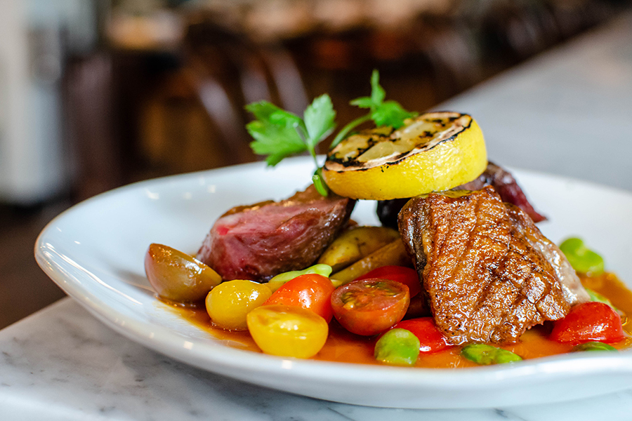 A French-style duck dish is topped with a charred lemon segment and sits in a thin reddish pool on a white plate.