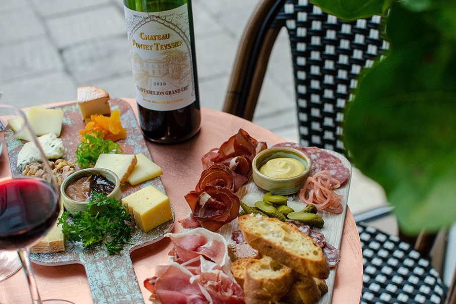 Charcuterie and cheese boards sit on a round copper table with a bottle and glass of red wine.