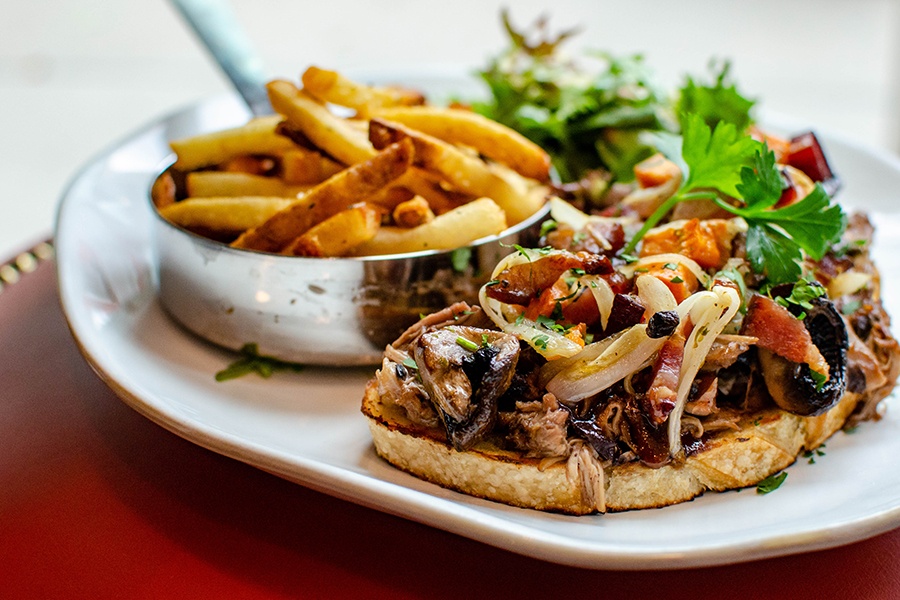 A thin piece of toast is covered with braised mushrooms and herbs, with a silver bowl of fries on the side.