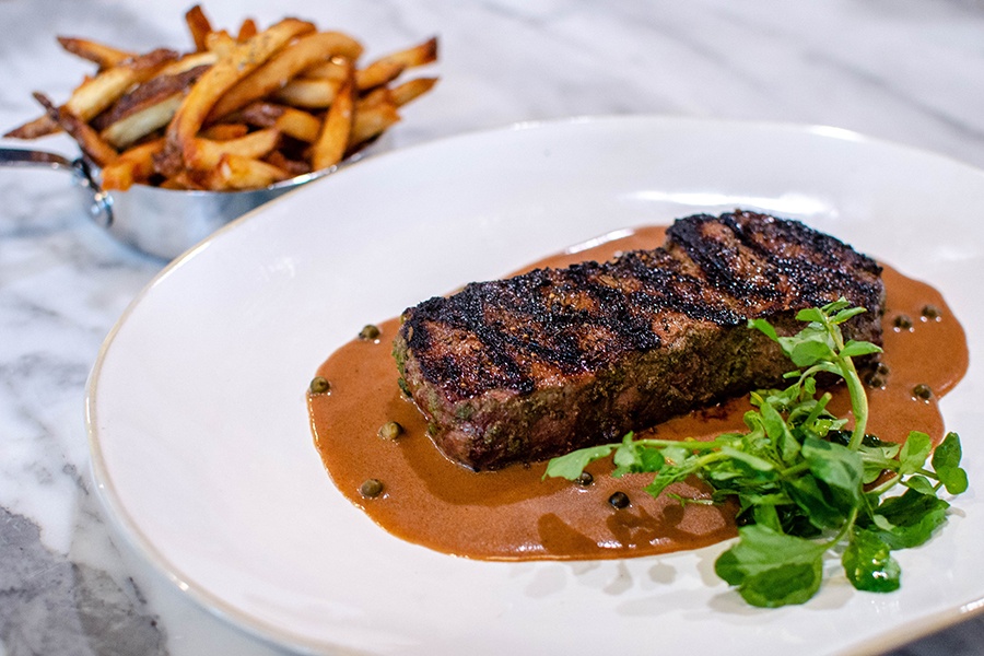A charred sirloin sits in a pool of a creamy light brown sauce with a side of arugula and some fries.