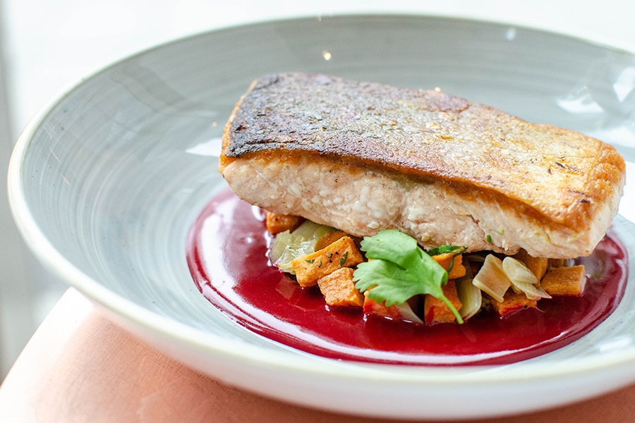 A salmon filet with crispy skin sits on a pile of chopped vegetables in a bright magenta pool of sauce.