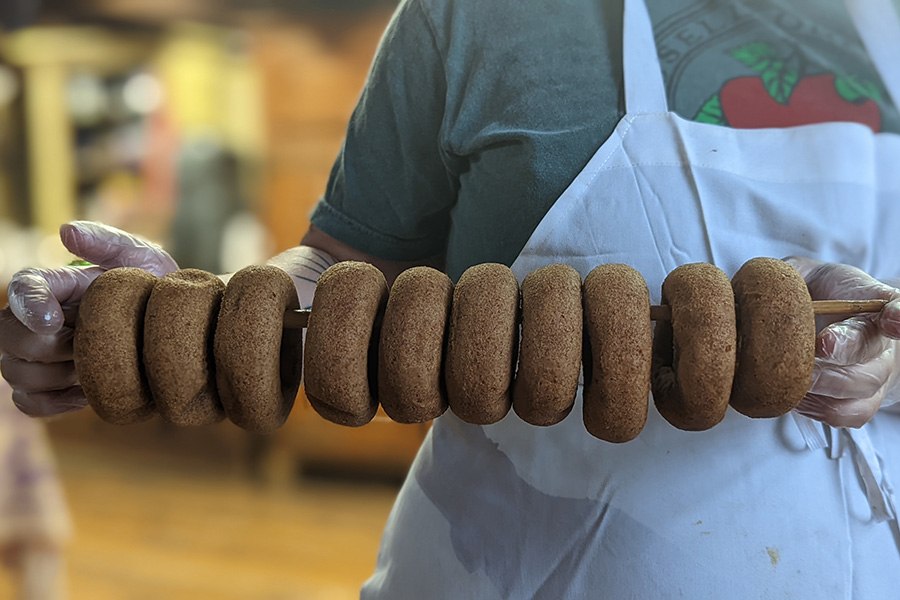 A person in a white apron and t-shirt with an apple design holds a wooden rod full of cider doughnuts.