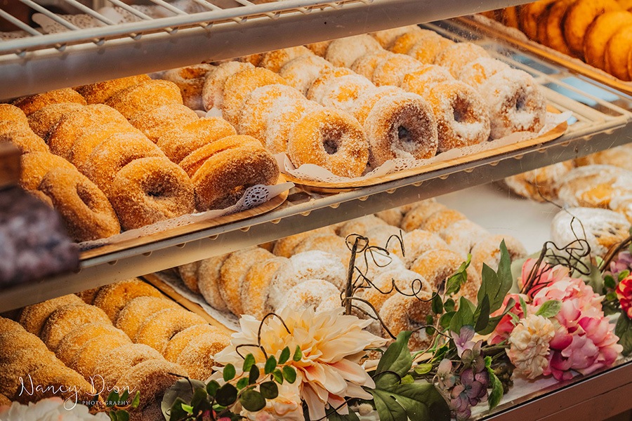 Rows of sugary cider doughnuts in a pastry case embellished with fresh flowers.