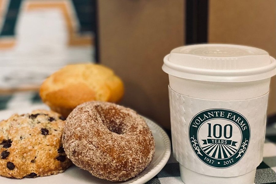 A sugary cider doughnut and other baked goods sit on a plate next to a paper cup of coffee with Volante Farms branding on the sleeve.