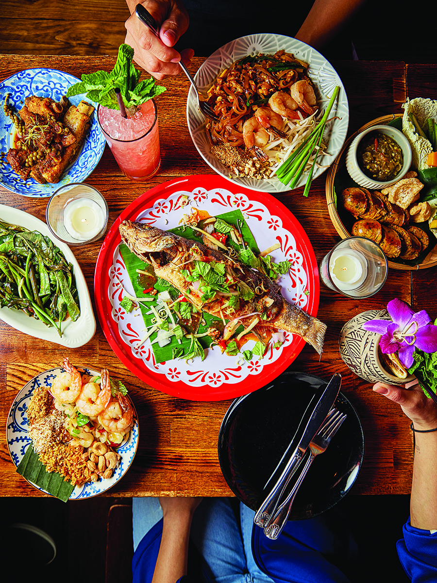 Overhead view of a wooden table full of colorful Thai dishes, including a whole fried fish as the centerpiece.