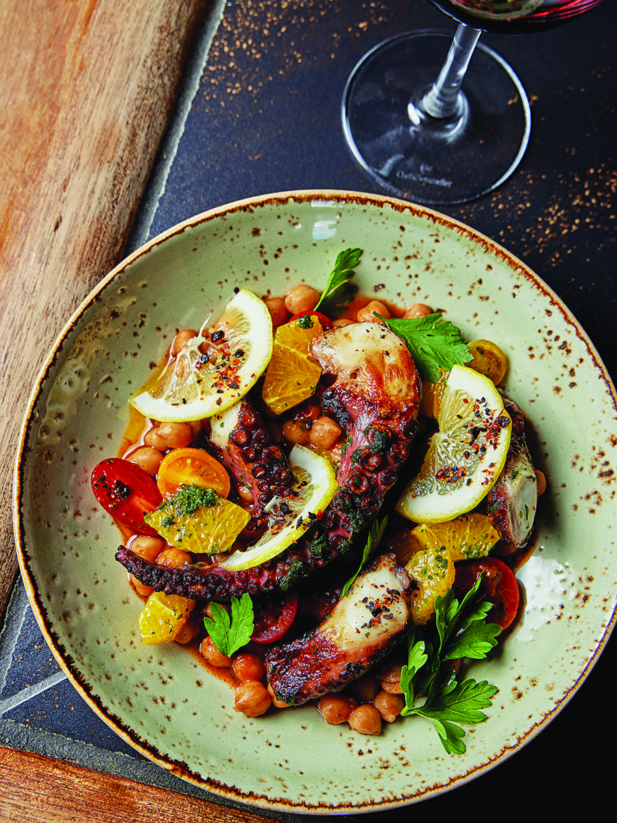 Overhead view of an octopus tendril garnishes with citrus slices, tomatoes, and chickpeas.