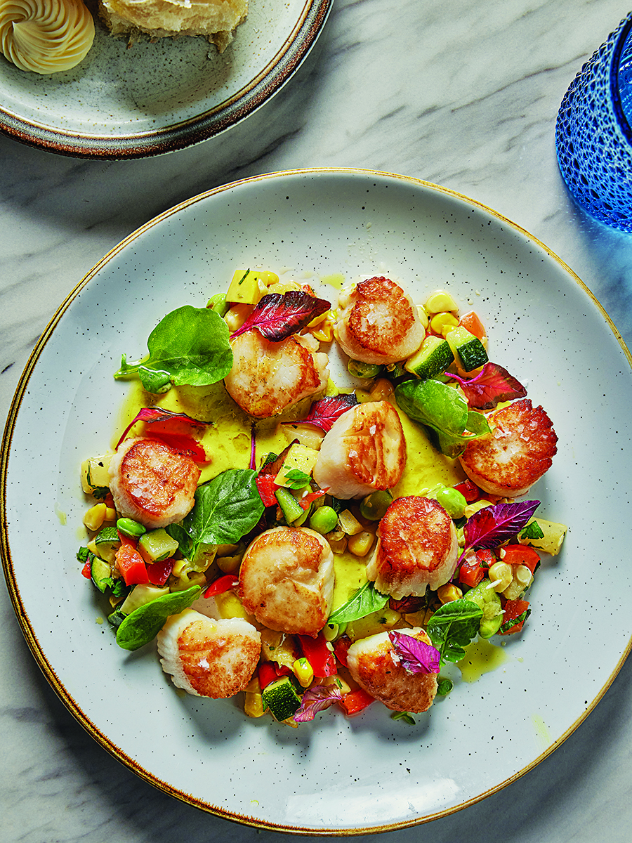 Overhead view of seared scallops on a bright yellow puree with lots of green veggies.