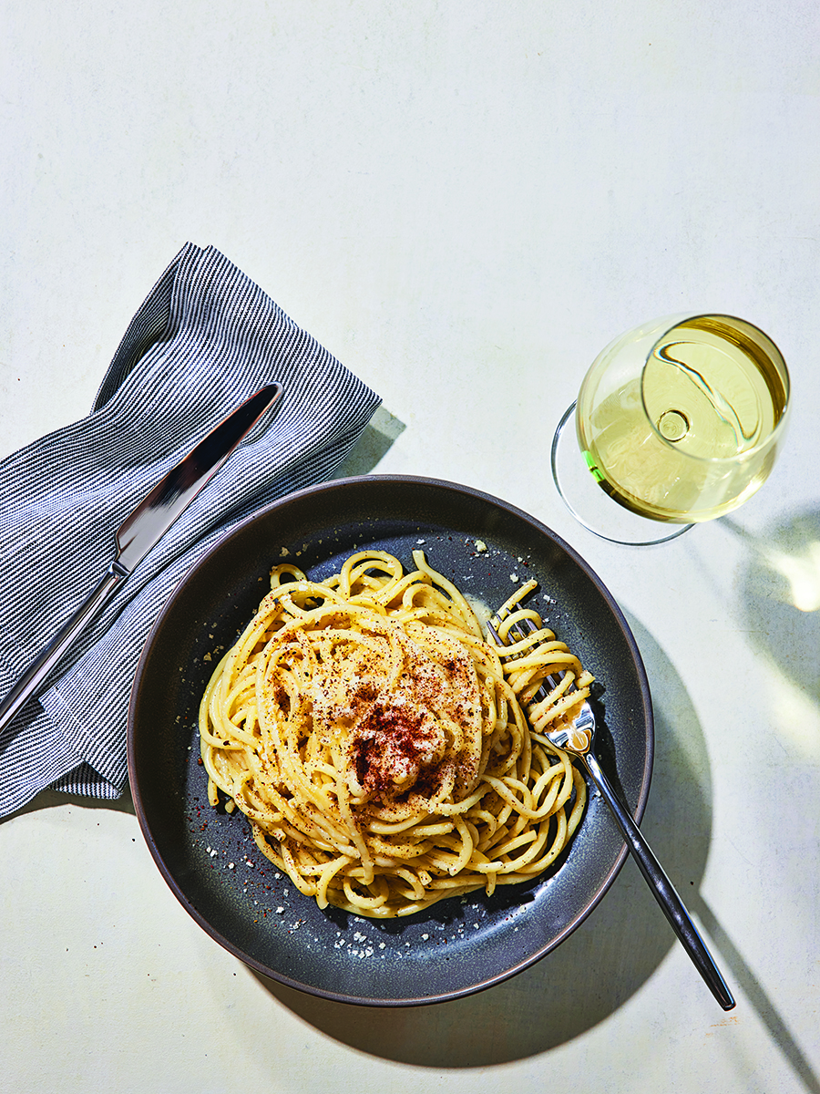 A pile of bucatini in a buttery sauce with cheese and grated pink pepper sits on a dark plate with a glass of white wine to the side.