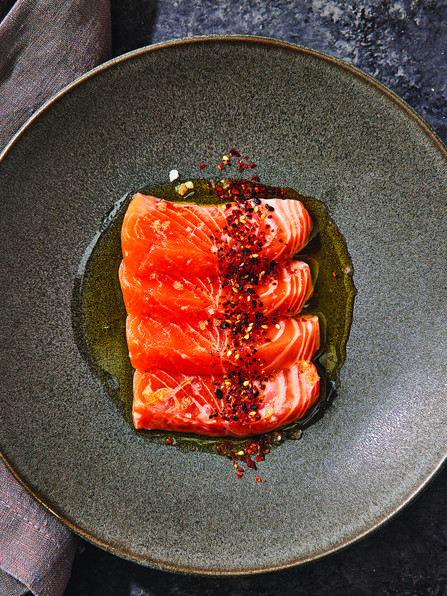Overhead view of a salmon crudo, with four plump slices lined up on a slate plate in a pool of olive oil, dusted with chili flakes.