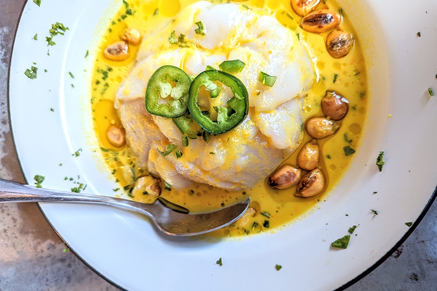 Slices of raw scallop are garnished with big toasted corn kernels and sliced green chili peppers, all sitting in a bright yellow broth.