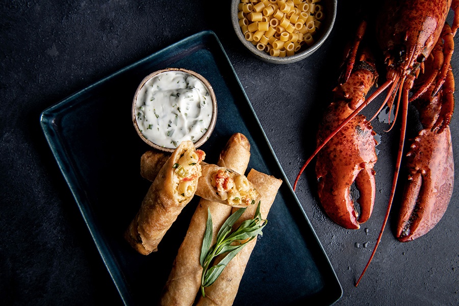 Spring rolls stuffed with lobster mac and cheese are displayed next to a bowl of uncooked pasta and a whole lobster.