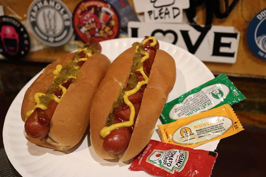 Two simple hot dogs are on a white paper plate, topped with squiggles of ketchup, mustard, and relish, displayed in front of a dive bar wall covered with stickers.