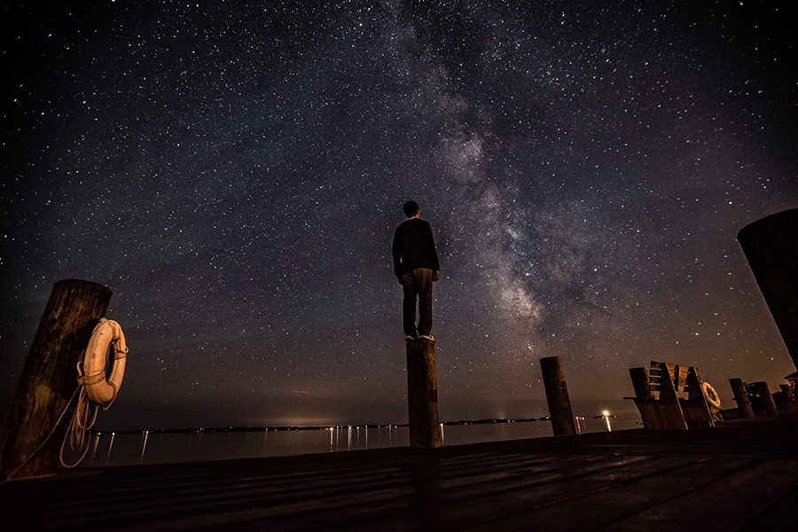 A man stands on a pier at night, back to the camera under a star-filled sky.