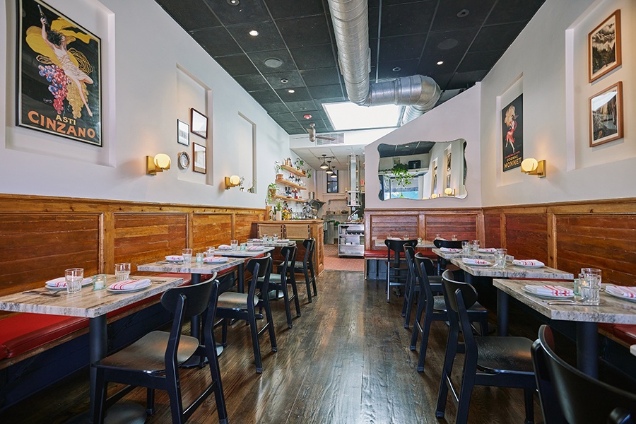 Interior view of a small, sun-lit restaurant with wood banquettes, red cushions, and vintage Italian art.